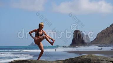 穿着比基尼<strong>站在</strong>一条腿<strong>上</strong>做瑜伽的年轻女孩<strong>站在</strong>海边的一块黑沙滩<strong>上</strong>